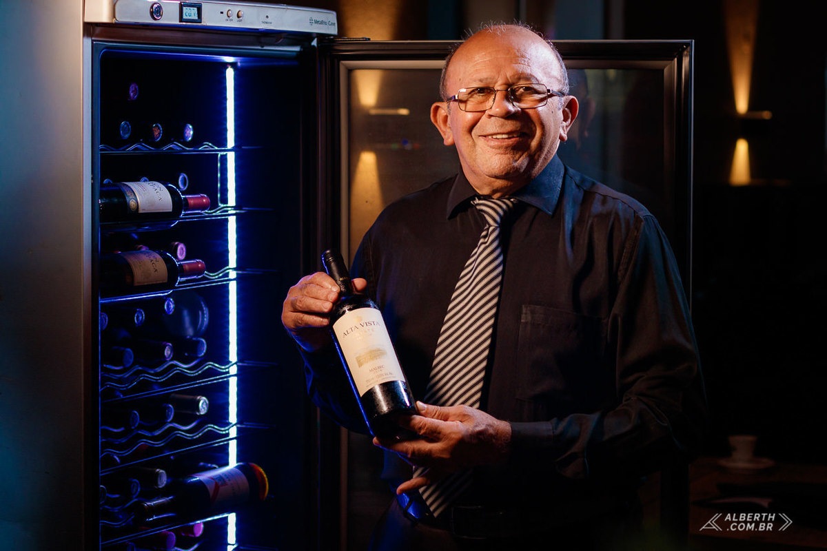 Retrato do senhor Otoniel Abílio da Costa, primeiro sommelier do Brasil, segurando um vinho com as duas mãos utilizando uma camisa social preta com uma gravata lpreto e branca listrada e com uma adega de vinhos ao seu lado