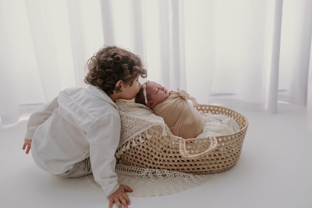 Irmão mais velho beijando o recém-nascido deitado em um moisés durante o ensaio newborn