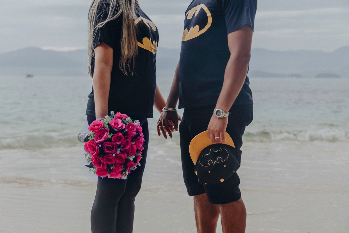 Casal apaixonado, vestidos com a camiseta do batman, um de frente para o outro com o mar de fundo, a noiva segurando o bouquet e o noivo segurando um boné do batman
