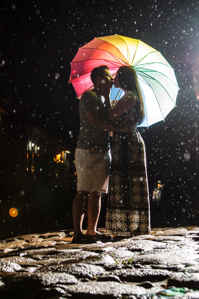 Casal apaixonado se beijando durante a noite enquanto seguram um guarda chuva colorido e gotas de chuva caem