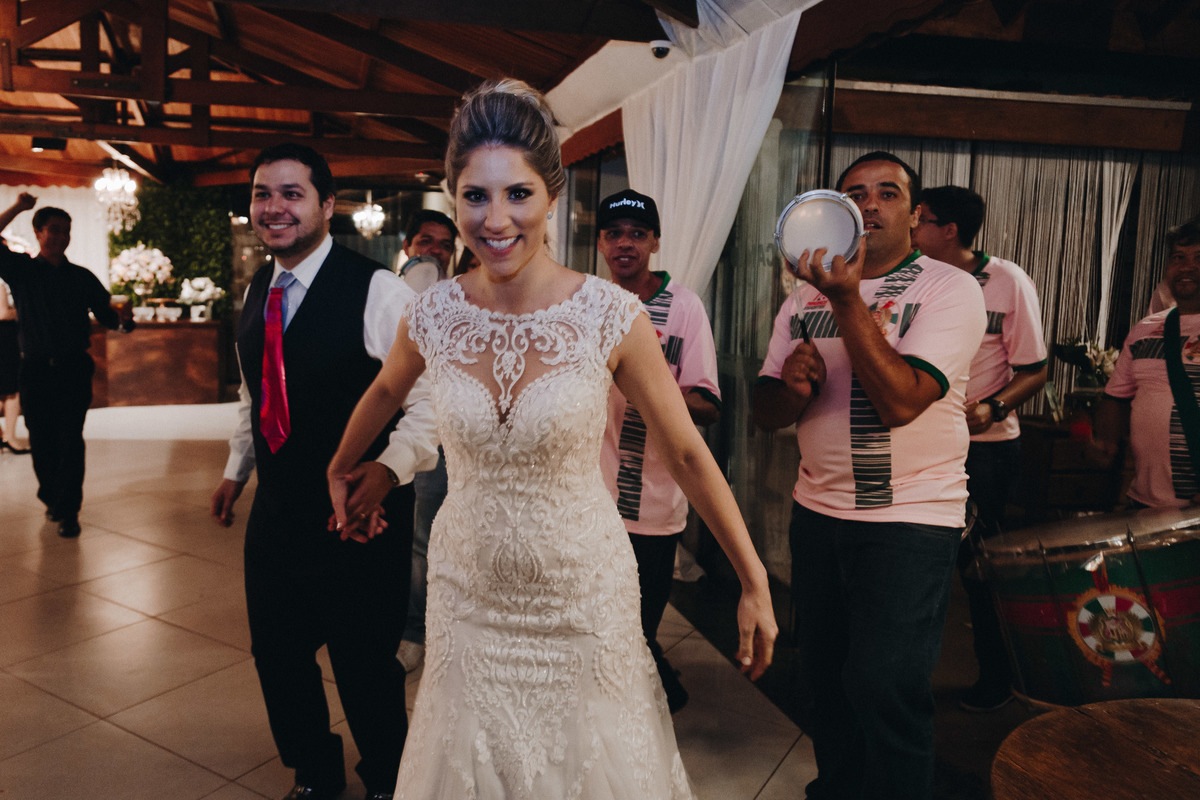 Noiva linda de mãos dadas com o noivo sorrindo para a frente com o batuque da escola de samba ao fundo