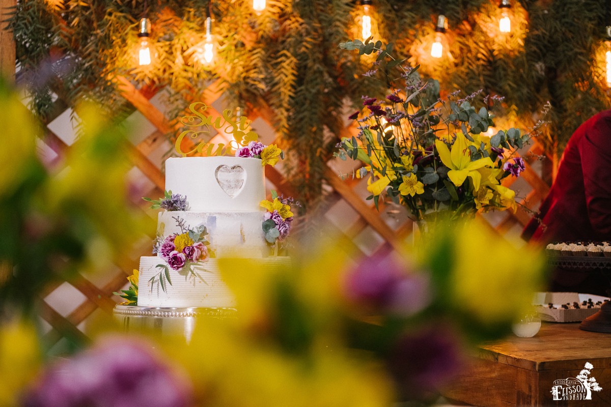 Bolo de casamento em decoração DIY com flores amarelas, roxas e folhagem verde, tudo rústico com madeira