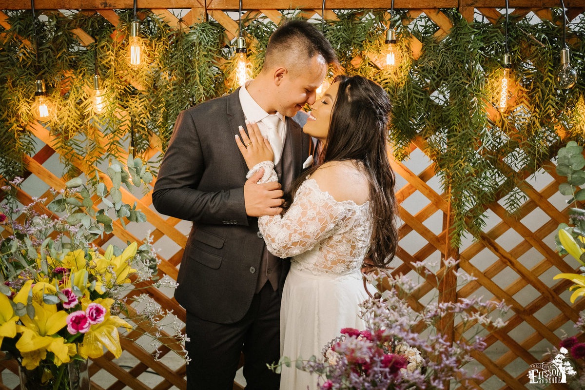 Casal abraçado em frente à decoração DIY com flores amarelas, roxas e folhagem verde, tudo rústico com madeira