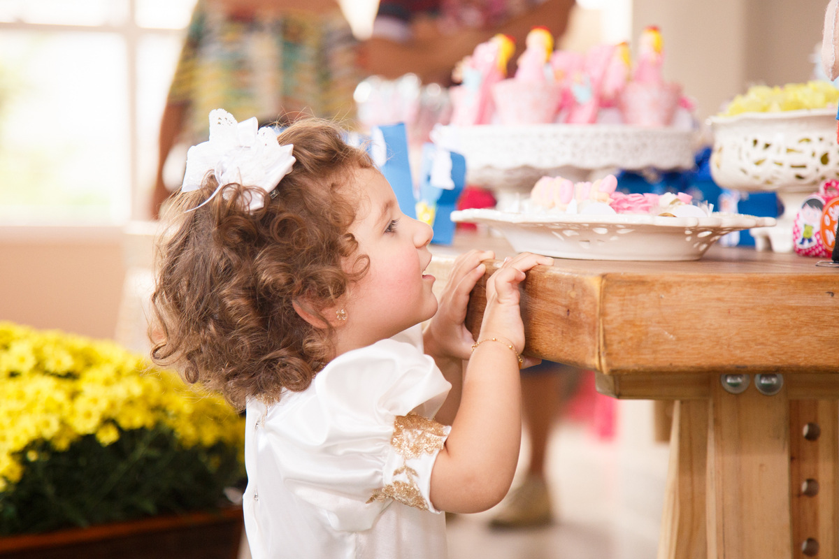 Criança de 2 anos em seu aniversário, na ponta dos pés olhando por cima da mesa do bolo.