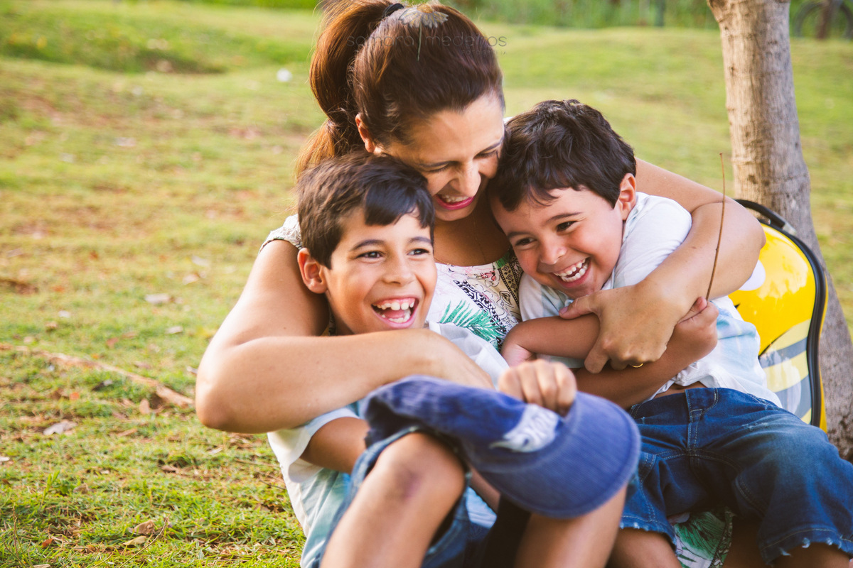 Mãe abraçando dois filhos e fazendo cócegas neles enquanto brincam em um ensaio fotográfico externo de família.