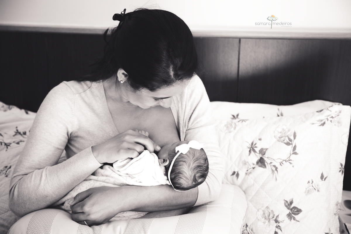 Foto em preto e branco de uma mãe sentada em sua cama amamentando sua filha recém nascida.