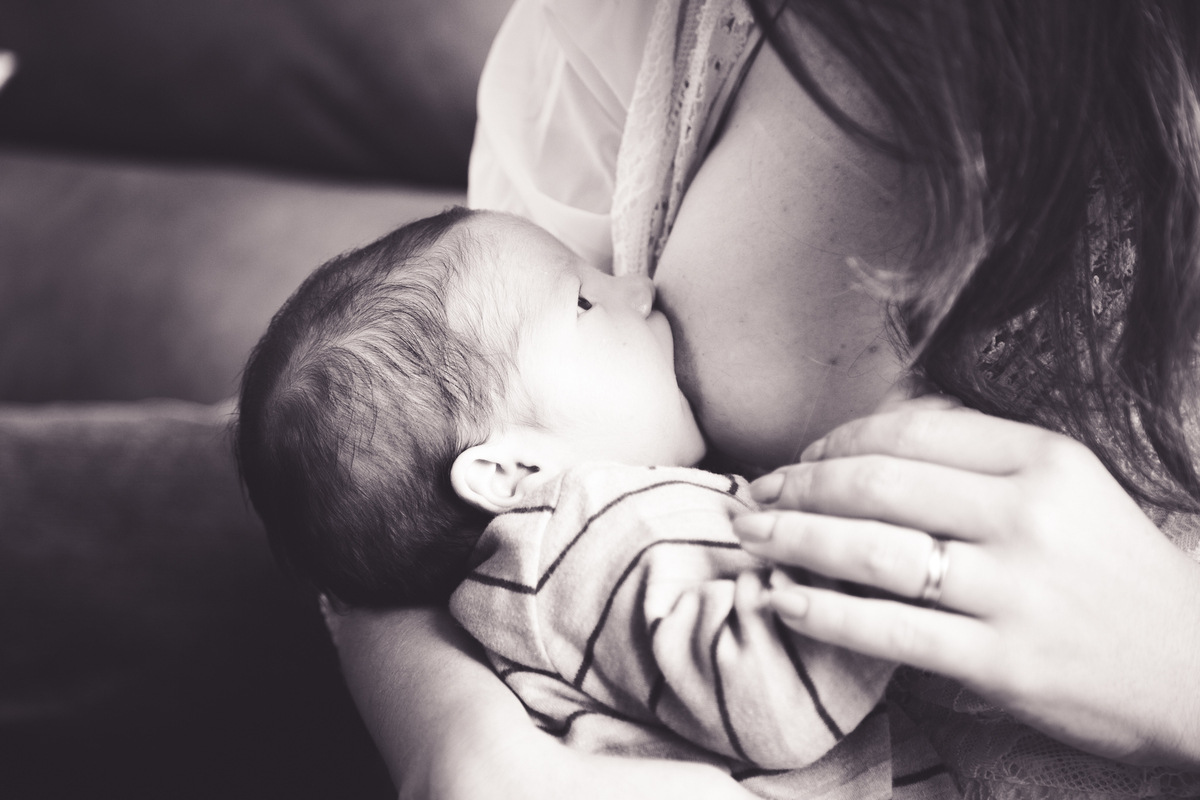 foto em preto e branco em close no bebê mamando, com os olhinhos abertos, olhando para a mãe, que segura sua mãozinha.