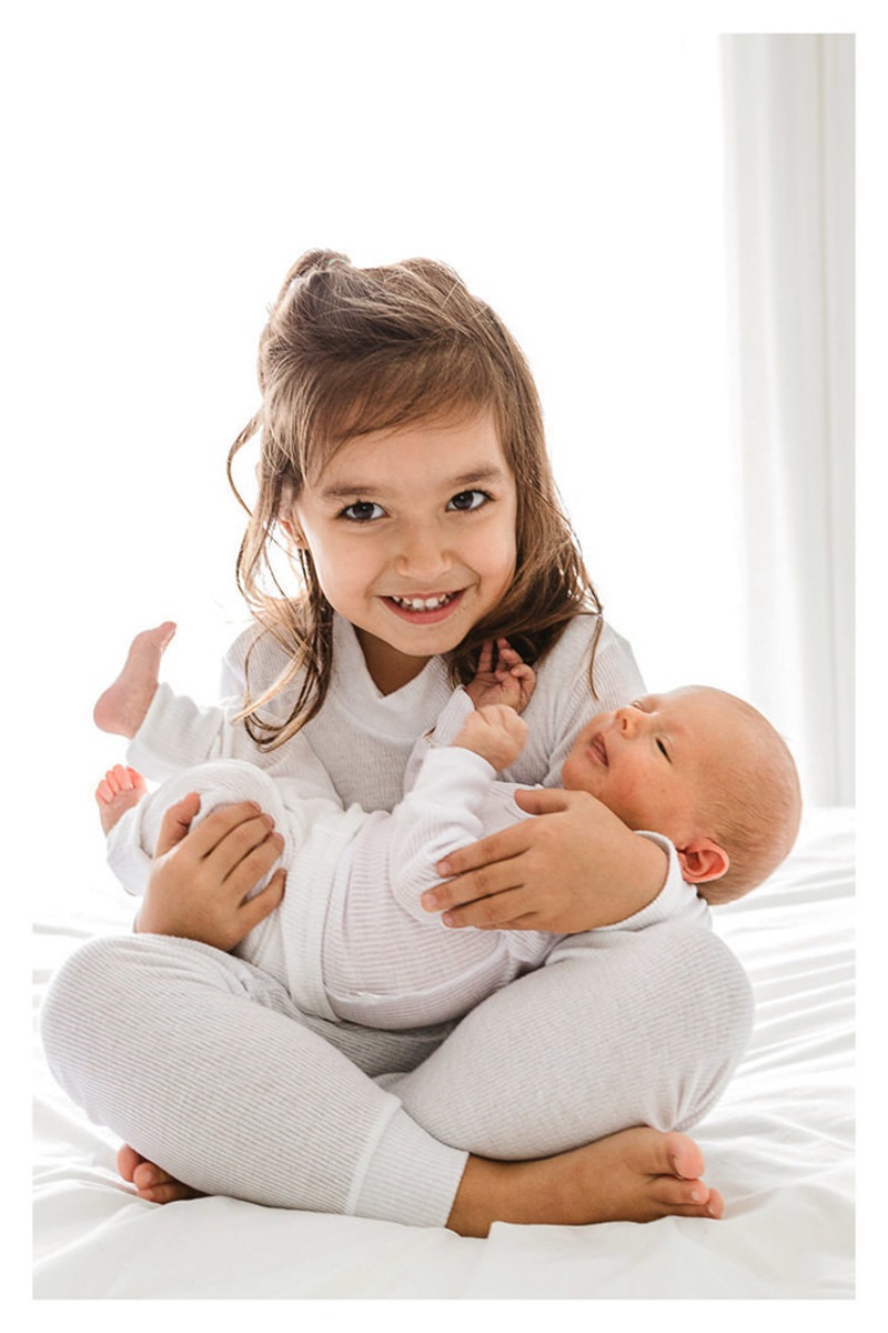 irmã sorrindo sentada na cama com bebê recém nascido no colo durante a sessão de fotos de newborn lifestyle em Curitiba, do fotógrafo infantil e de família Rapha Luna