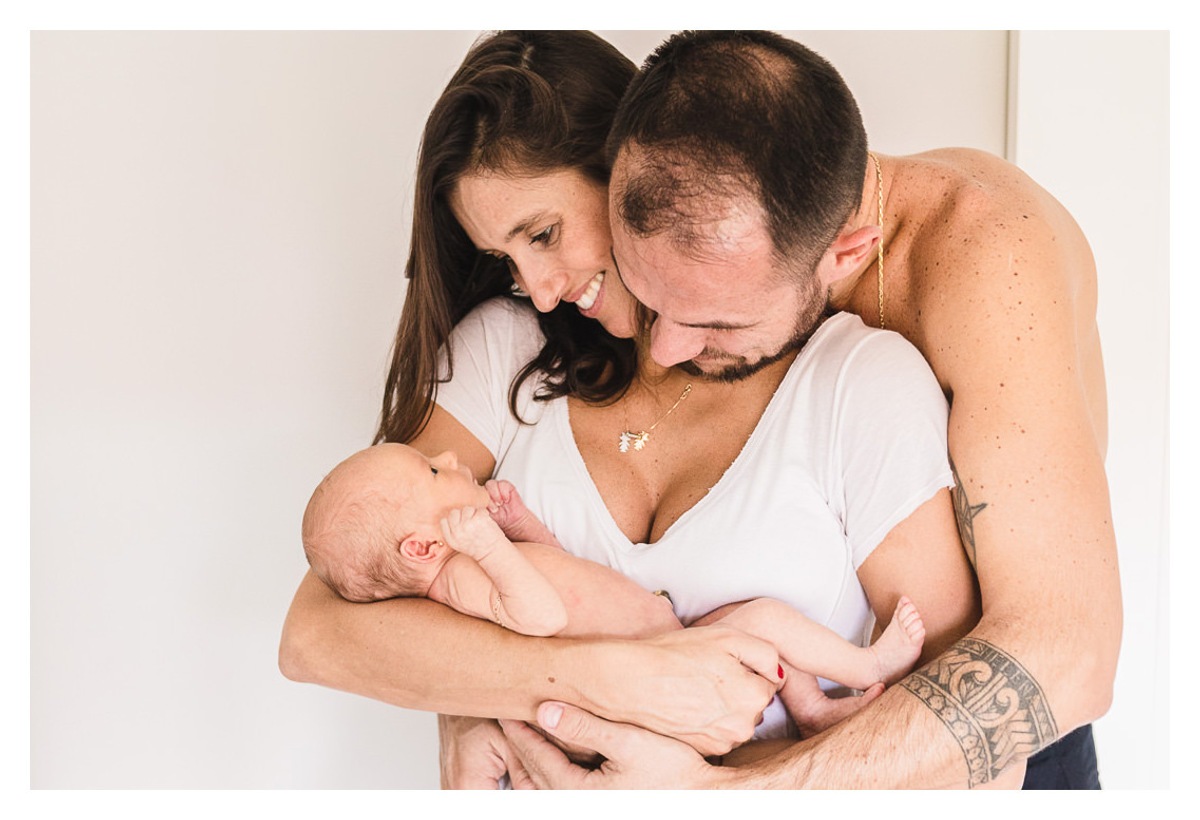 Mãe com bebê recém nascido, newborn no colo, sendo abraçada pelo pai em casa, durante a sessão de fotos de newborn lifestyle em Curitiba, do fotógrafo infantil e de família Rapha Luna