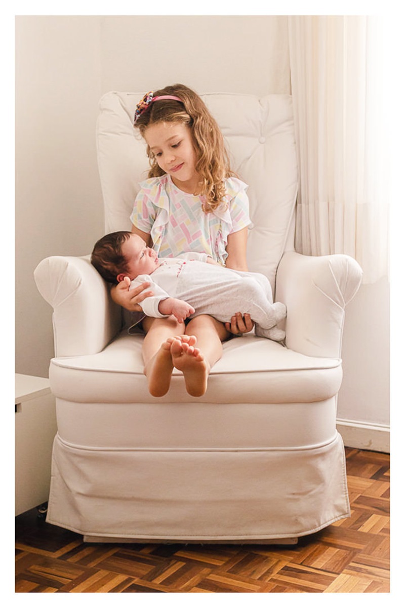 irmã sentada na poltrona de amamentação com bebê recém nascido no colo, durante a sessão de fotos de newborn lifestyle em Curitiba, do fotógrafo infantil e de família Rapha Luna