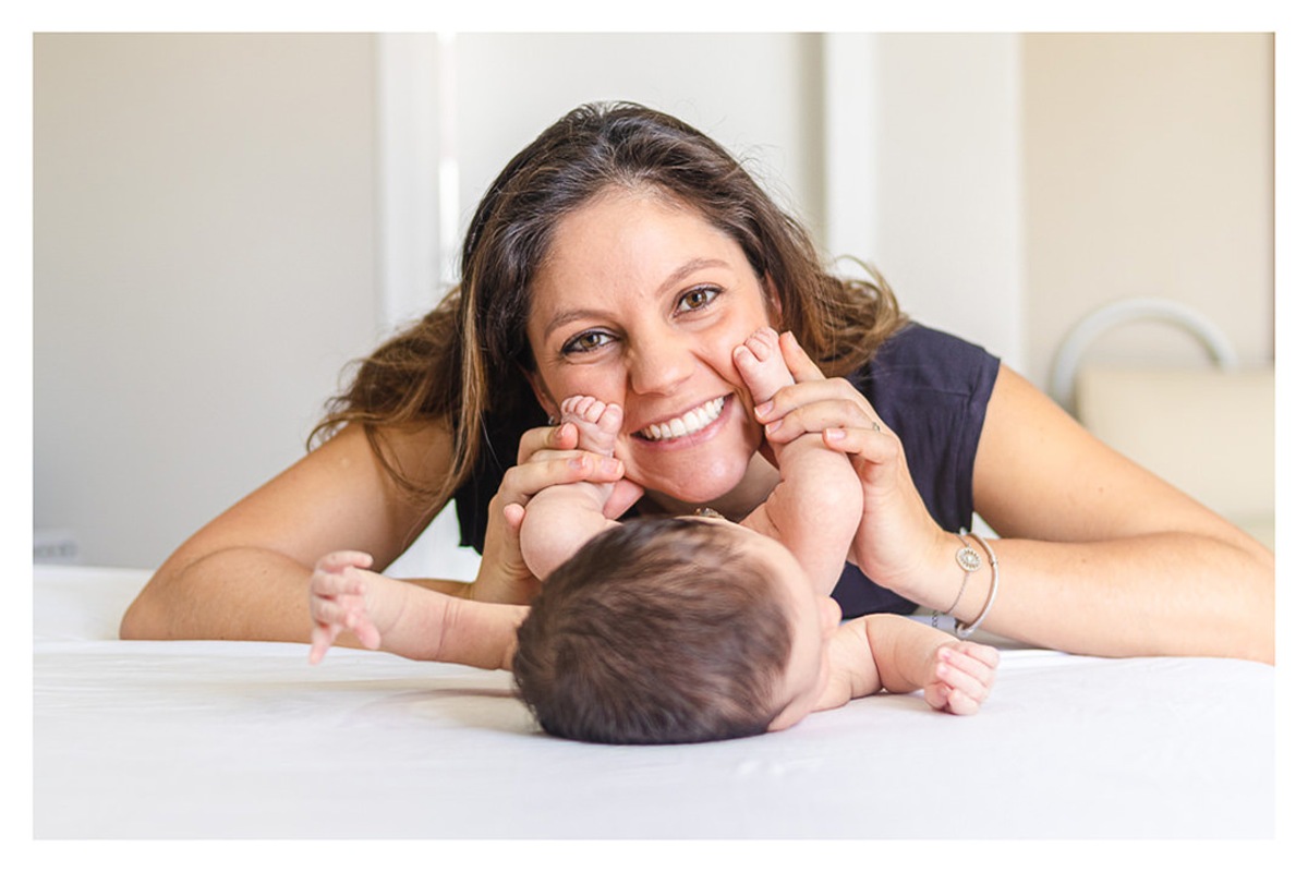 Mãe deitada na cama, sorrindo e brincando com os pés do bebê recém nascido, durante a sessão de fotos de newborn lifestyle em Curitiba, do fotógrafo infantil e de família Rapha Luna