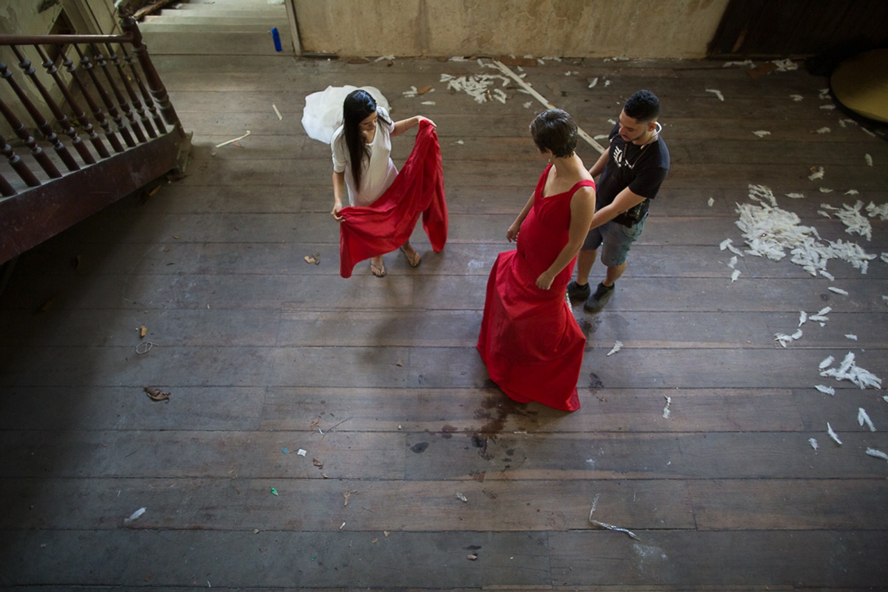 Cena de bastidores de uma produção artística, com três pessoas em um salão amplo de piso de madeira. Uma delas veste um vestido vermelho enquanto as outras ajustam a peça e posicionam o tecido. O ambiente apresenta marcas de uso e elementos espalhados pelo chão, sugerindo um espaço criativo em processo.