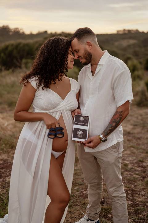 Casal mostra ecografia e sapatinhos de bebé durante sessão fotográfica de gravidez.'