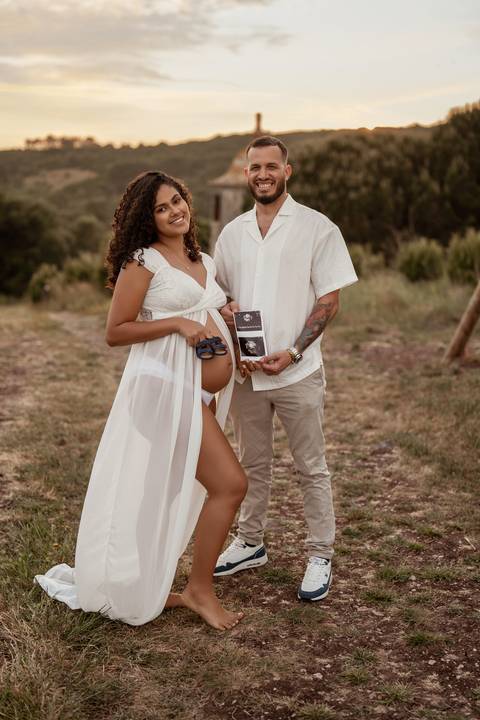 Pai abraça mulher grávida e segura barriga durante sessão fotográfica ao pôr do sol.'