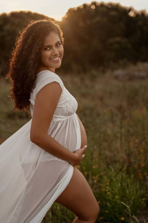 Mulher grávida sorridente com vestido branco durante sessão fotográfica de gravidez em exterior ao pôr do sol em Lisboa.'