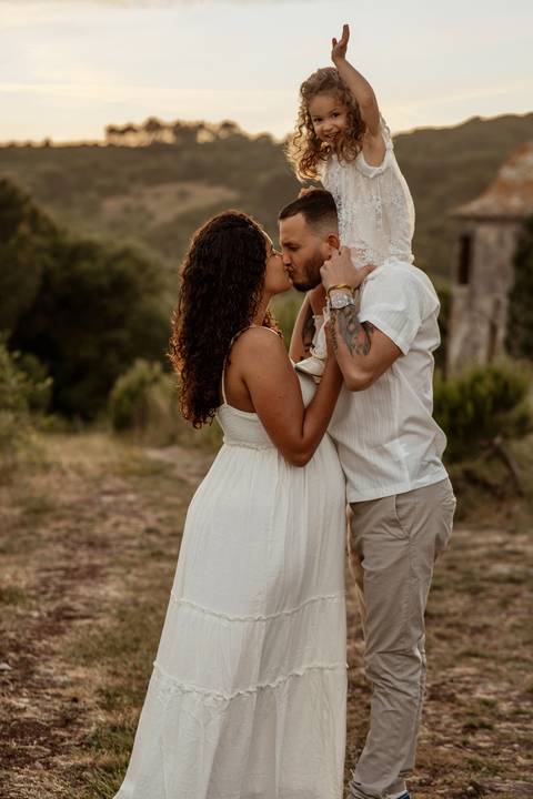 Casal grávido com filha pequena durante sessão fotográfica de família ao ar livre.'