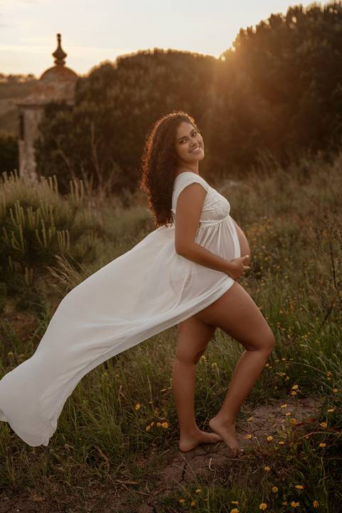 Mulher grávida sorridente com vestido branco durante sessão fotográfica de gravidez em exterior ao pôr do sol em Lisboa.'
