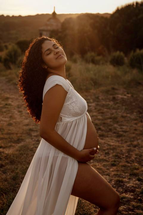 Mulher grávida sorridente com vestido branco durante sessão fotográfica de gravidez em exterior ao pôr do sol em Lisboa.'