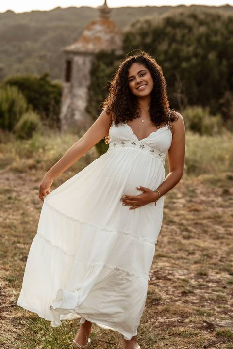 Mulher grávida sorridente com vestido branco durante sessão fotográfica de gravidez em exterior ao pôr do sol em Lisboa.'