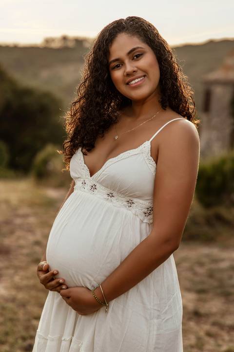 Mulher grávida sorridente com vestido branco durante sessão fotográfica de gravidez em exterior ao pôr do sol em Lisboa.'