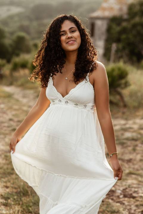 Mulher grávida sorridente com vestido branco durante sessão fotográfica de gravidez em exterior ao pôr do sol em Lisboa.'