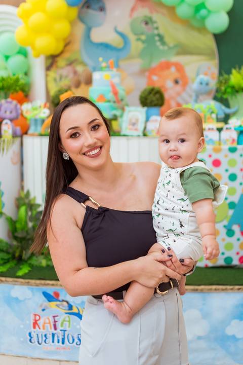 Mãe segurando Gustavo no colo ao lado do bolo de aniversário, com decoração de dinossauros ao fundo.'