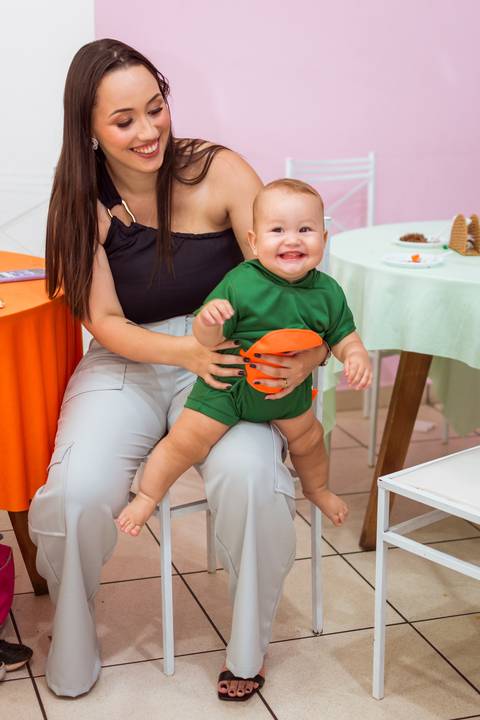 Gustavo sorrindo com sua fantasia verde de dinossauro enquanto a mãe o ajuda a ficar em pé durante sua festa de 1 aninho.'