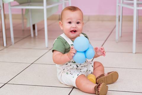 Gustavo sentado e sorrindo enquanto brinca com um brinquedo azul durante sua festa de 1 ano.'