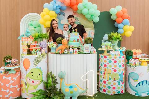 Família do Gustavo reunida atrás da mesa decorada com dinossauros para celebrar seu primeiro aniversário.'