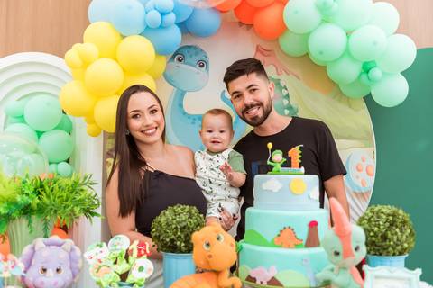 Pais e Gustavo sorrindo juntos em frente à mesa do bolo durante seu aniversário de 1 ano com tema Dinossauros em Tatuí.'