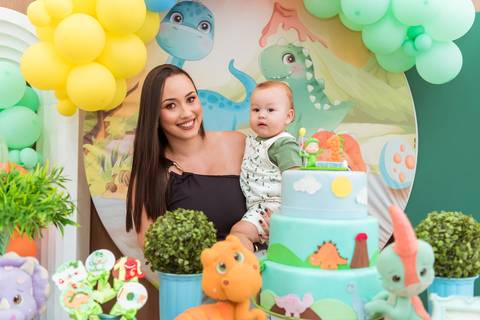 Gustavo no colo da mãe ao lado do bolo de aniversário decorado com dinossauros, em um registro especial do seu primeiro aninho.'