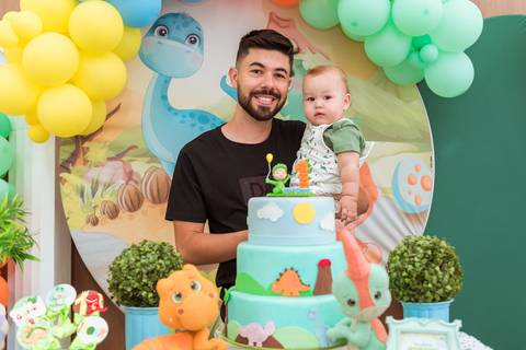 Pai sorrindo com Gustavo no colo atrás do bolo temático de dinossauros durante sua festa de 1 ano no Rafa Eventos em Tatuí.'