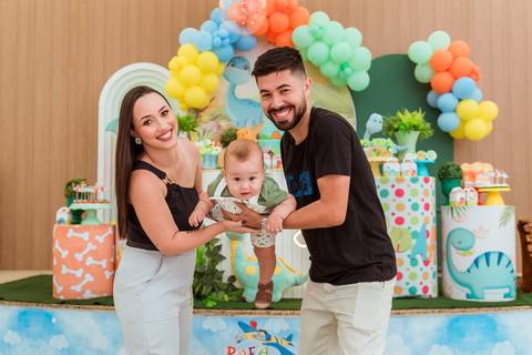 Pai levantando Gustavo no colo enquanto a mãe acompanha o momento com carinho durante a festa infantil.'