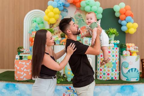 Pai levantando Gustavo no colo enquanto a mãe acompanha o momento com carinho durante a festa infantil.'