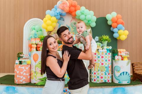 Pai levantando Gustavo no colo enquanto a mãe acompanha o momento com carinho durante a festa infantil.'