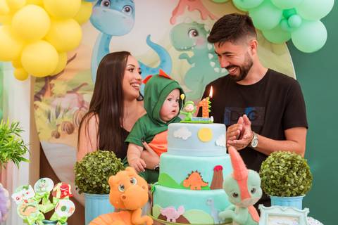 Pais do Gustavo sorrindo e interagindo com o filho ao lado do bolo de dinossauros durante a festa de 1 aninho em Tatuí.'