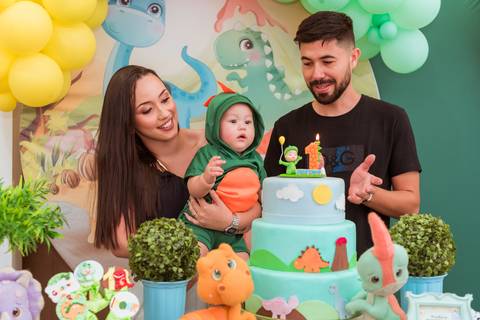 Gustavo fantasiado de dinossauro no colo da mãe, acompanhado do pai em frente ao bolo de seu primeiro aniversário.'