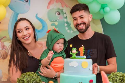 Família reunida em um momento de alegria, com Gustavo vestido de dinossauro junto aos pais na hora do parabéns.'