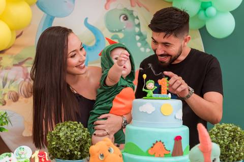 Gustavo sorrindo no colo da mãe enquanto o pai brinca com ele ao lado do bolo temático de dinossauros.'