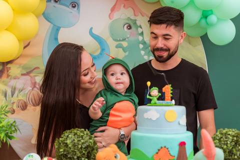 Gustavo fantasiado de dinossauro no colo da mãe, acompanhado pelo pai em frente ao bolo de seu aniversário de 1 ano.'