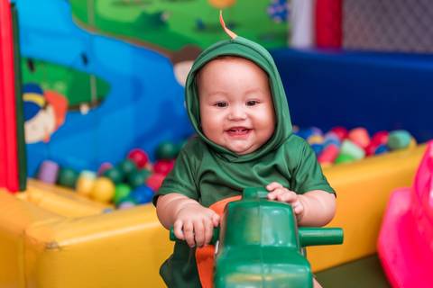 Gustavo sorrindo enquanto brinca em um dinossauro de balanço verde, vestido com sua fantasia temática na festa de aniversário.'