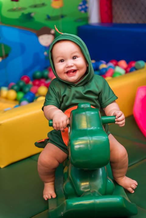 Detalhe de Gustavo segurando o brinquedo de dinossauro verde, com um sorriso de alegria durante a festa de 1 ano.'