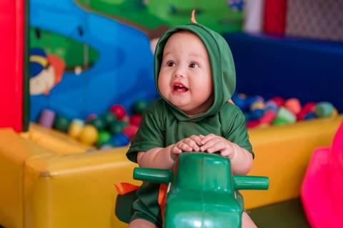 Gustavo vestido de dinossauro olhando para o lado enquanto brinca em um balanço verde no espaço infantil da festa.'