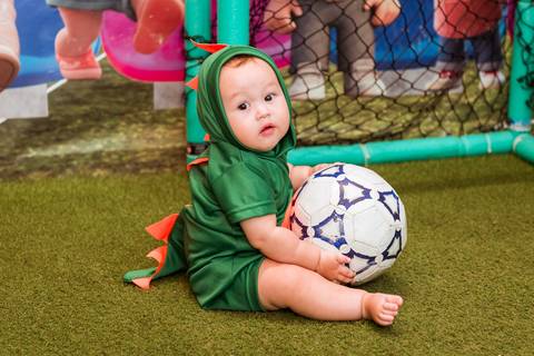 Gustavo fantasiado de dinossauro sentado no gramado sintético, segurando uma bola de futebol durante sua festa de 1 ano.'