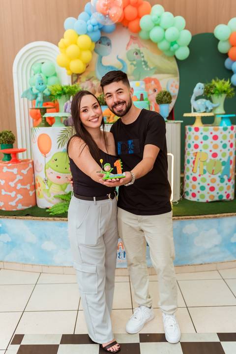 Pais do Gustavo segurando juntos uma lembrança de dinossauro personalizada com o número 1 durante a festa de aniversário.'