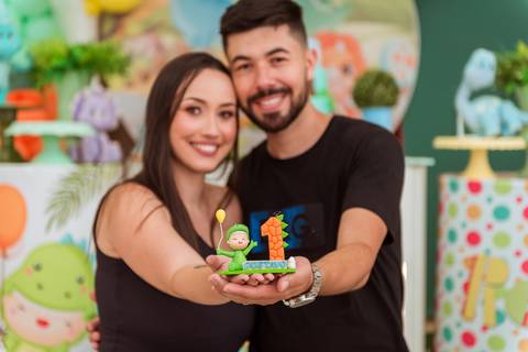 Casal sorrindo e apresentando uma decoração de dinossauro com o número 1, celebrando o primeiro aninho do filho Gustavo.'