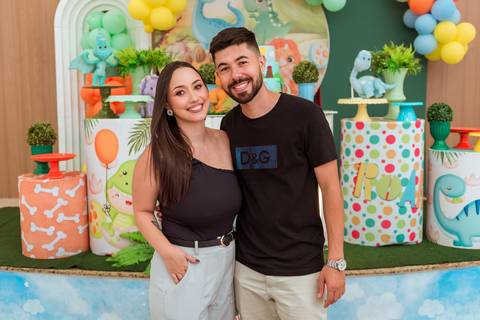Pais do Gustavo posando juntos em frente à decoração temática de dinossauros durante a festa infantil no Rafa Eventos em Tatuí.'