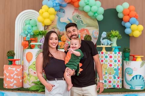 Gustavo no colo do pai, acompanhado pela mãe, em uma fotografia de família durante seu aniversário de 1 ano com tema Dinossauros.'
