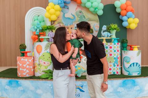 Gustavo no colo do pai, acompanhado pela mãe, em uma fotografia de família durante seu aniversário de 1 ano com tema Dinossauros.'