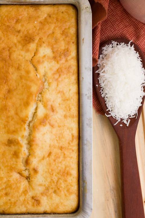 bolo de coco em uma assadeira de alumínio feito artesanalmente, ao lado vemos uma colher de pau com lascas de coco fresco'